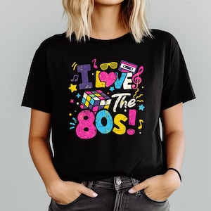 Puede incluir: Camiseta negra con un gr&aacute;fico colorido que dice "I Love The 80s!". El dise&ntilde;o incluye un cubo de Rubik, notas musicales, gafas de sol, una cinta de cassette y estrellas. El texto y los gr&aacute;ficos son de colores brillantes y retro.