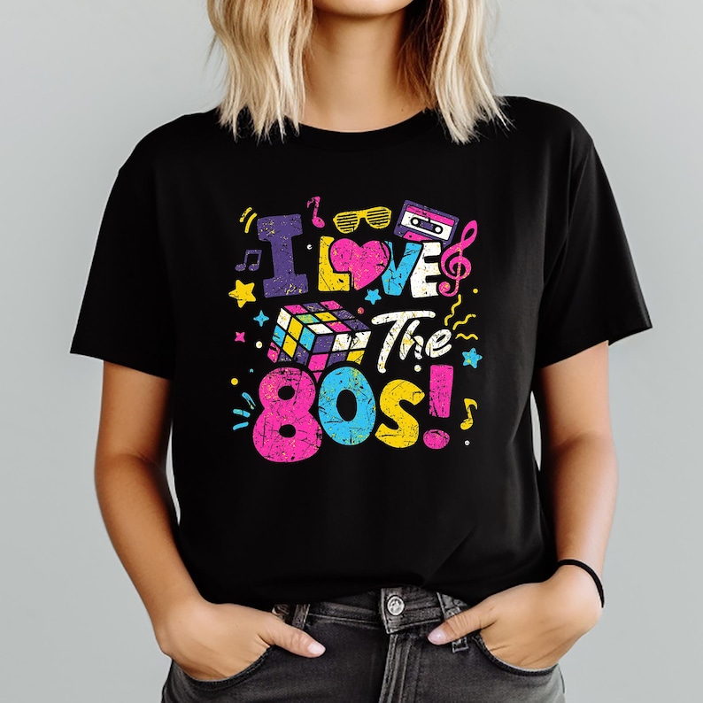 Puede incluir: Camiseta negra con un gr&aacute;fico colorido que dice "I Love The 80s!". El dise&ntilde;o incluye un cubo de Rubik, notas musicales, gafas de sol, una cinta de cassette y estrellas. El texto y los gr&aacute;ficos son de colores brillantes y retro.