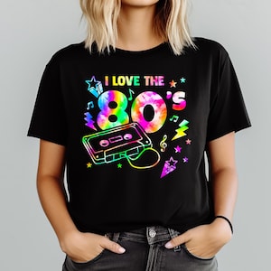 Puede incluir: Camiseta negra con un dise&ntilde;o gr&aacute;fico colorido que dice "I LOVE THE 80'S". El dise&ntilde;o incluye un "80's" estilo tie-dye, una cinta de cassette, notas musicales, estrellas y rayos.