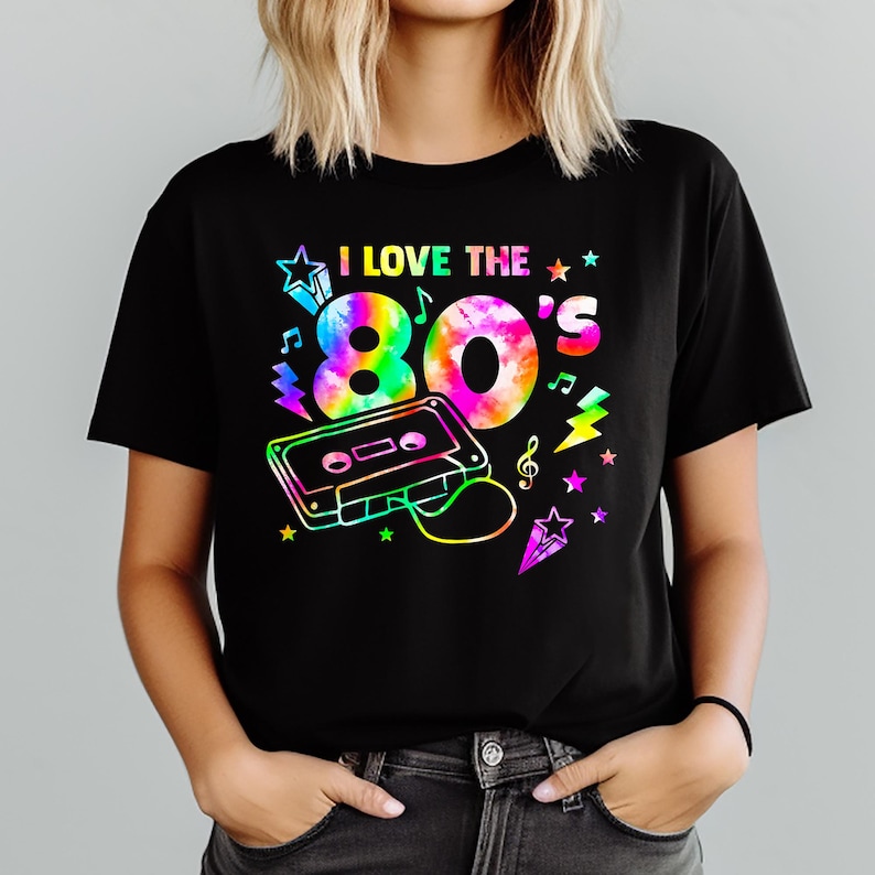 Puede incluir: Camiseta negra con un dise&ntilde;o gr&aacute;fico colorido que dice "I LOVE THE 80'S". El dise&ntilde;o incluye un "80's" estilo tie-dye, una cinta de cassette, notas musicales, estrellas y rayos.