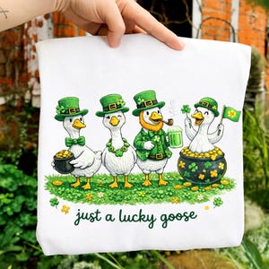 Puede incluir: Bolso de mano blanco con cuatro gansos de dibujos animados vestidos con atuendos del Día de San Patricio. Cada ganso lleva un sombrero verde y está rodeado de monedas de oro y tréboles. El texto "just a lucky goose" está escrito en verde.