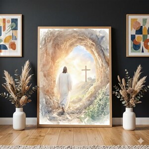 Puede incluir: Una impresión en acuarela enmarcada representa una figura con una túnica blanca caminando hacia una cruz de madera. La escena está ambientada dentro de una abertura de cueva, bañada por la luz del sol. La obra está enmarcada en madera clara y expuesta en una pared oscura.