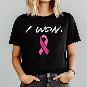 May include: Black t-shirt featuring the white text "I WON." above a pink ribbon. The t-shirt is a crew neck style. The person is wearing the t-shirt.