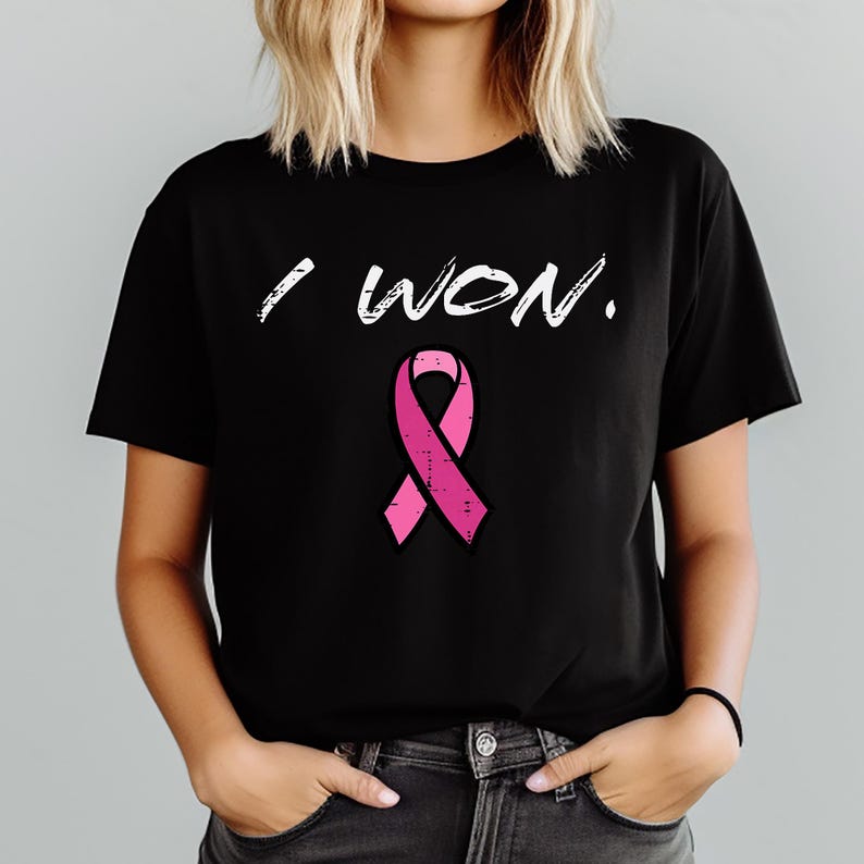 May include: Black t-shirt featuring the white text "I WON." above a pink ribbon. The t-shirt is a crew neck style. The person is wearing the t-shirt.