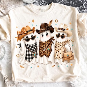 Puede incluir: Una camiseta de color crema con un gráfico de tres fantasmas que llevan sombreros de vaquero y accesorios de estilo western. Los fantasmas están adornados con estampados de leopardo, cuadros vichy en blanco y negro, lunares y gafas de sol.