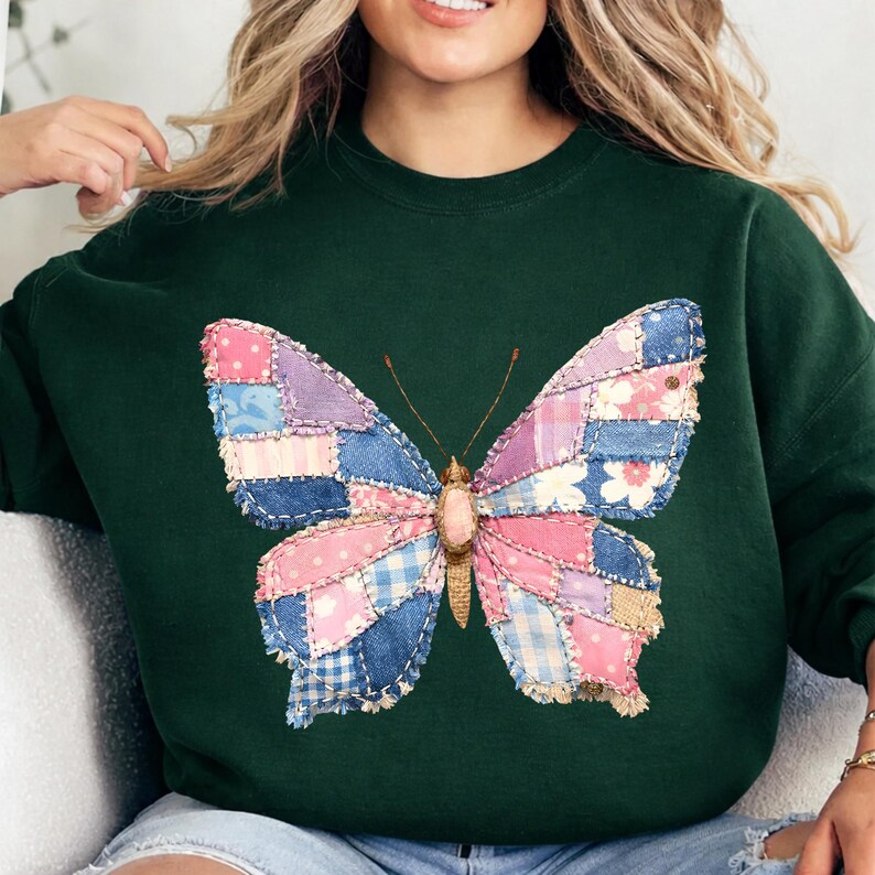 Pode incluir: Camisola verde escuro com um grande desenho de borboleta. As asas da borboleta s&atilde;o feitas de tecido patchwork em tons de rosa, azul e roxo, com estampas florais e xadrez. A borboleta tem um corpo e antenas bege.