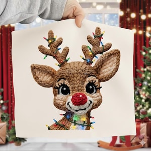 Puede incluir: Ilustración de un reno tejido con una nariz roja, adornado con luces navideñas de colores en sus cuernos y alrededor de su cuello. El reno tiene ojos grandes y expresivos y está sobre un fondo crema, evocando un tema festivo navideño.