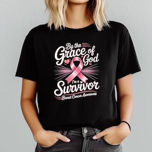 May include: Black t-shirt featuring a pink ribbon and the words "By the Grace of God I'm a Survivor" in white. The shirt also includes the text "Breast Cancer Awareness".