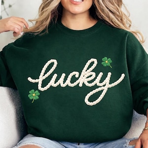 Puede incluir: Sudadera verde oscuro con la palabra "lucky" bordada en blanco, con letras cursivas. Dos diseños de tréboles verdes están bordados en la sudadera. La sudadera es de cuello redondo.