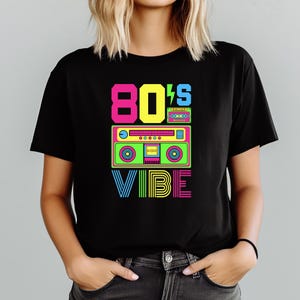 Puede incluir: Camiseta negra con un gr&aacute;fico colorido de los a&ntilde;os 80. El dise&ntilde;o presenta el texto "80's Vibe" con un radiocasete y un rayo. El texto y el radiocasete son de colores ne&oacute;n brillantes.