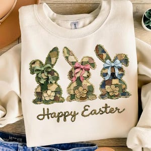 Puede incluir: Sudadera crema con tres conejos aplicados con patrones de patchwork, cada uno adornado con un lazo y una flor. El texto "Happy Easter" está bordado debajo de los conejos, lo que lo convierte en una prenda festiva con temática de Pascua.