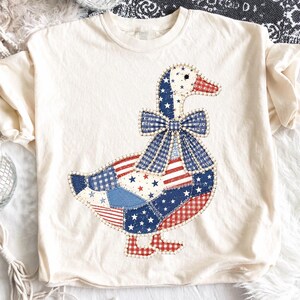 Puede incluir: Una camiseta de manga corta color crema con un gran gráfico de un ganso. El ganso está diseñado con un patrón de patchwork de telas rojas, blancas y azules, incluyendo estrellas, rayas y cuadros vichy, y lleva un gran lazo de cuadros azules.