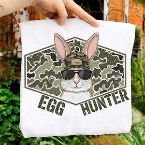 Puede incluir: Bolso de mano blanco con un conejo de dibujos animados con gafas de sol y un sombrero de camuflaje. El conejo está dentro de un hexágono de camuflaje verde y gris con las palabras "EGG HUNTER" impresas debajo.