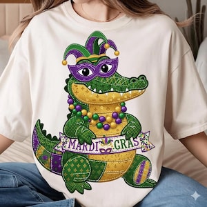May include: A cream-coloured t-shirt with a cartoon alligator wearing a purple mask and jester hat. The alligator has a beaded necklace and holds a banner reading "MARDI GRAS". The design uses green, purple, and gold colours, perfect for a festive occasion.