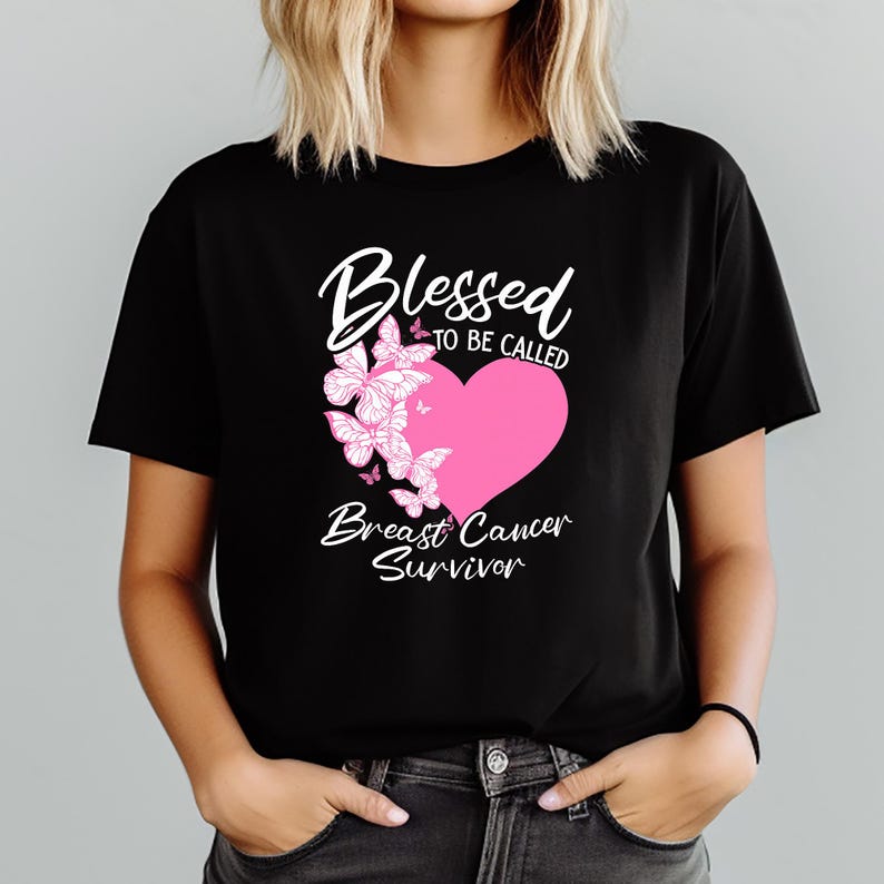 May include: Black t-shirt featuring a pink heart and butterfly design. White script reads "Blessed to be Called Breast Cancer Survivor". The short-sleeved shirt is worn by a person.