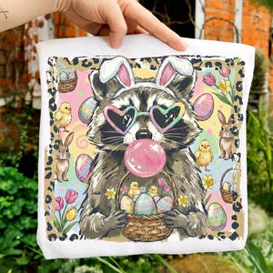Puede incluir: Cojín cuadrado blanco con un mapache con gafas de sol en forma de corazón y orejas de conejo, soplando una burbuja rosa. El diseño incluye huevos de Pascua, pollitos, conejos y flores. Un borde de estampado de leopardo enmarca la imagen, creando un tema festivo de Pascua.