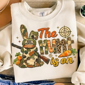 Puede incluir: Sudadera de color crema con un gráfico colorido. El gráfico presenta un conejo de camuflaje con un sombrero que dice "Egg Hunter" y un rifle. También se muestra el texto "The Hunt is on".