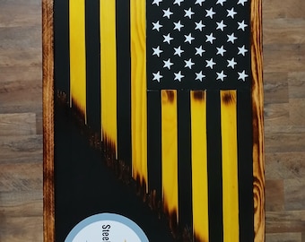 May include: Wooden American flag with a Steelers logo. The flag features yellow stripes, a black background, and white stars. The flag is framed with a dark brown wooden border. The Steelers logo is a circle with a blue, yellow, and red emblem.