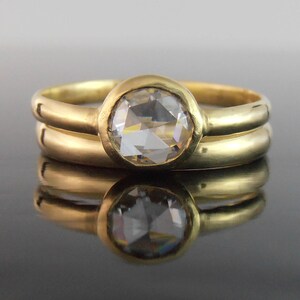 May include: A gold ring featuring a large, clear, round gemstone in a bezel setting, paired with a plain gold band. The jewellery is presented on a reflective surface.