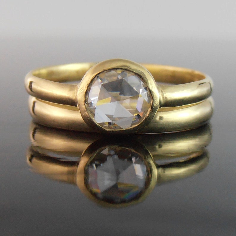 May include: A gold ring featuring a large, clear, round gemstone in a bezel setting, paired with a plain gold band. The jewellery is presented on a reflective surface.