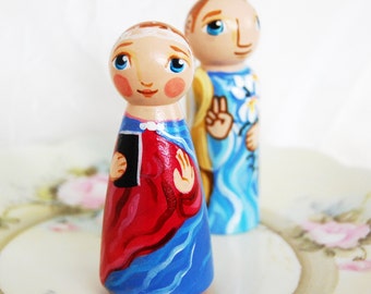 Puede incluir: Dos muñecas de madera pintadas a mano, una con una túnica roja y azul que sostiene un libro, la otra con una túnica azul con una flor blanca.