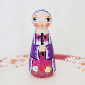 May include: A hand-painted wooden peg doll depicting a saintly figure. The doll wears a purple robe with white trim and holds a dark cross. The base of the doll is pink and decorated with painted flowers. The doll has blue eyes and a blue head covering.