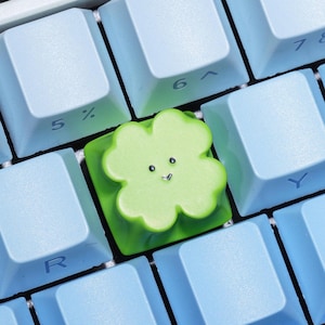 May include: A custom green clover-shaped keycap with a smiling face, set among light blue keyboard keys. The keycap is a unique accessory for mechanical keyboards, adding a touch of personality.