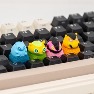 May include: Four colourful, animal-shaped keycaps sit on a black keyboard. The keycaps are blue, yellow, pink, and orange, each with unique features. The keyboard has black and off-white keys.