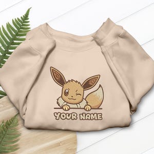 May include: A beige crewneck sweatshirt featuring an embroidered graphic of a winking, light brown creature with large ears and a fluffy tail, resting its paws on a horizontal line above the text "YOUR NAME" in brown block letters.