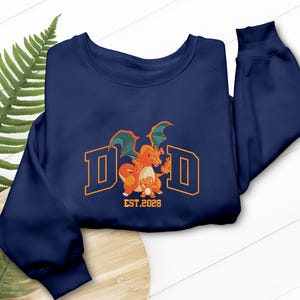May include: Navy blue sweatshirt with the word "DAD" in orange block letters, featuring a cartoon dragon graphic. The graphic includes the text "EST.2026". The sweatshirt is displayed on a wooden surface with a fern.