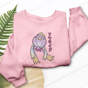 May include: A light pink sweatshirt featuring an embroidered cartoon character with a purple body, cream-colored hat, and yellow appendages. Japanese text is printed vertically on the right side of the design.
