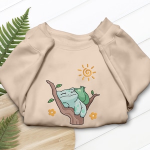 Op de afbeelding: Beige sweatshirt met een geborduurd ontwerp van een cartoon Bulbasaur en een kleiner groen wezen dat op een tak rust. Het ontwerp bevat een gele zon en bloemen. De sweatshirt heeft lange mouwen en een ronde hals.