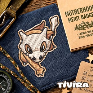 May include: A dark blue fabric patch featuring an embroidered Cubone character with a white skull mask and brown body. The patch is displayed alongside a tan card labelled MOTHERHOOD MERIT BADGE, a green braided cord, and a compass on a wooden surface.