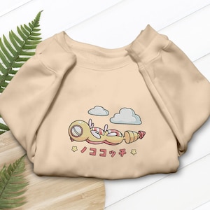 May include: A beige sweatshirt featuring a cartoon character with a yellow body, red accents, and a striped tail, lying under two cartoon clouds. Japanese text is below the character. The sweatshirt is made of a soft material.
