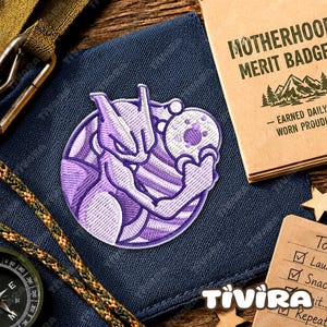 May include: A circular embroidered patch featuring a purple and white stylized character with pointed ears and a tail, holding a spherical object. The patch is displayed on a dark blue fabric surface next to a tan booklet labeled Motherhood Merit Badge.