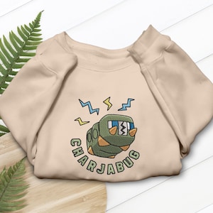 Op de afbeelding: Een beige sweatshirt met een geborduurd cartoon rupsontwerp en de tekst "CHARJABUG". De rups is groen en oranje met blauwe en gele bliksemschichten erboven. De sweatshirt is op een houten oppervlak gevouwen.