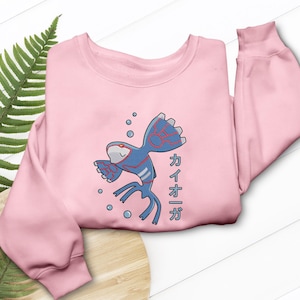 May include: A light pink sweatshirt featuring a blue and red cartoon fish design with Japanese text. The fish has a white belly and is surrounded by blue bubbles. The sweatshirt has long sleeves and a crew neck.