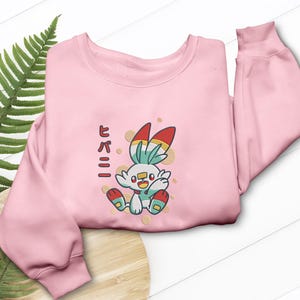 May include: A pale pink sweatshirt with a cartoon rabbit character in red, yellow, and blue. The design includes gold-coloured dots and Japanese text on the left side. A casual, comfortable garment.