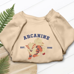 Könnte beinhalten: Beiges Sweatshirt mit gesticktem Design eines Comic-Caniden in Orange, Rot und Creme. Das Wort "Arcanine" ist über dem Caniden gewölbt, darunter "Est. 1996".