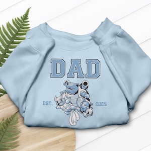 May include: Light blue sweatshirt with the word "DAD" in a blue, block letter design. Below the word "DAD" is a graphic of three cartoon characters. The text "EST. 2025" is below the graphic.