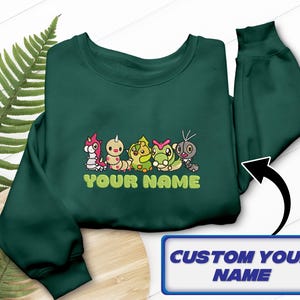 Könnte beinhalten: Dunkelgrünes Sweatshirt mit einer Stickerei von bunten Cartoon-Kreaturen. Darunter ist der Text "YOUR NAME" grün gestickt. Eine blau-weiße Box unten zeigt "CUSTOM YOUR NAME".