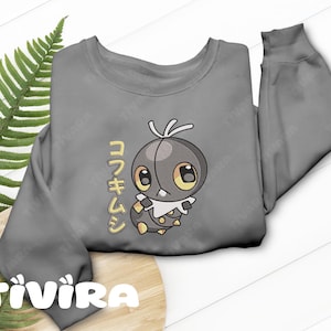 Op de afbeelding: Een grijs sweatshirt met lange mouwen en een afbeelding van een cartooninsect met grote ogen, een witte kraag en gele accenten. Links van het personage staat verticale Japanse tekst in het geel, gecentreerd op de voorzijde van het kledingstuk.