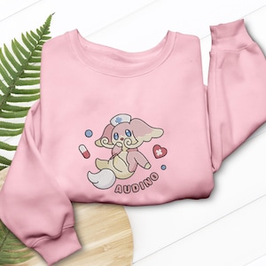 May include: A pale pink sweatshirt with an embroidered cartoon character wearing a nurse's hat, a heart, and a pill. The character is in pink, cream, and blue. The word "AUDINO" is written in a circle below the character.