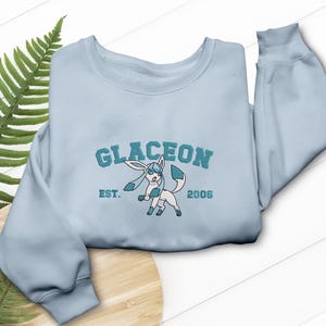 May include: A pale blue sweatshirt featuring teal embroidery of the word "GLACEON", a cartoon character, and the text "EST. 2006". The sweatshirt has a round neck and long sleeves.