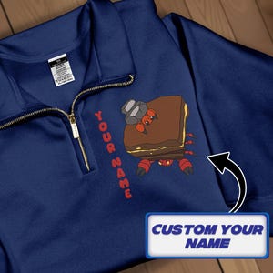 May include: Navy blue quarter-zip pullover with a gold zipper. The shirt features a cartoon crab design with a rock hat and the text "YOUR NAME" in red. A text box says "CUSTOM YOUR NAME".