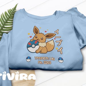 Könnte beinhalten: Ein hellblaues Sweatshirt mit einer Stickerei des braunen Eevee-Charakters, der einen rot-weiß-blauen Pokéball hält. Das Design enthält gelbe Sterne, rote Herzen, japanischen Text und die Aufschrift: TRAINER: Lily No. 2268.