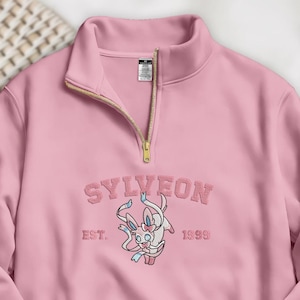 Könnte beinhalten: Ein hellrosa Sweatshirt mit Viertelreißverschluss und goldenem Reißverschluss. Das Sweatshirt zeigt das gestickte Wort "SYLVEON" über einem Cartoon-Charakter, darunter "EST. 1999". Das Sweatshirt ist aus weichem Material.