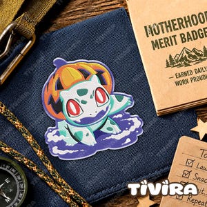 May include: An embroidered patch featuring a light green creature with red eyes wearing an orange pumpkin on its head, surrounded by a purple cloud-like base. The patch is displayed on a dark blue fabric surface next to a tan booklet.