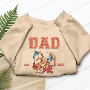 May include: A beige sweatshirt featuring the word "DAD" in a red, retro font. Below the text are three cartoon characters and the text "EST. 2025". The sweatshirt is folded on a wooden surface.