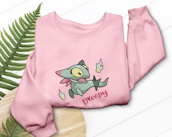 May include: A light pink sweatshirt featuring an embroidered cartoon character. The character is green with a pink scarf and the name "Dreepy" in pink script. The sweatshirt has long sleeves and a crew neck.